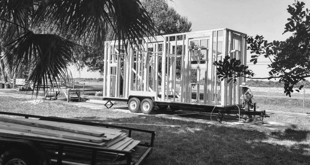 Black-and-white photo of a framed tiny house on a trailer.